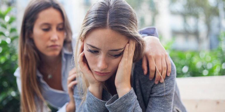 Concerned woman comforting distressed woman experiencing signs of prescription drug addiction while sitting on a park bench together